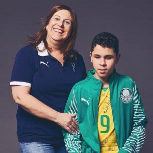 Una mujer abrazando a su hijo en un estudio de fotografía. Ella tiene una camiseta azul del club de fútbol Palmeiras de Brasil y su hijo, quien es ciego, un polerón verde del mismo club.