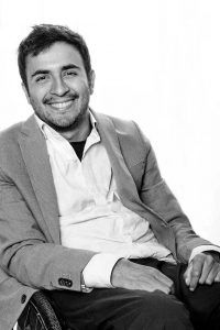 Un hombre en silla de ruedas y vestido con terno y camisa sonríe a la cámara. La foto está en blanco y negro.