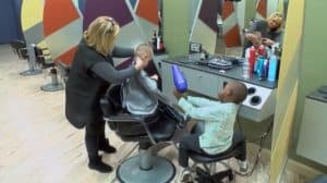 Dos niños de diferentes razas en una peluquería mientras a uno de ellos le están cortando el pelo.
