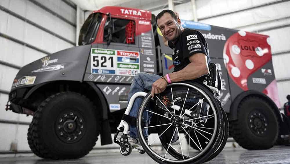 El piloto Albert Llovera sonriendo en su silla de ruedas y atrás se ve el enorme camión de carreras que conduce.