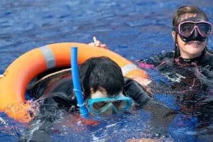 una mujer y un joven en el agua con traje de buceo, y ella le sostiene un flotador.