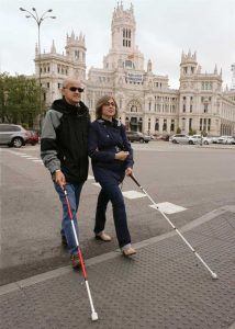 Una persona sordociega con su bastón blanco con rojo, camina por una vereda junto a una persona ciega que va con su bastón blanco.