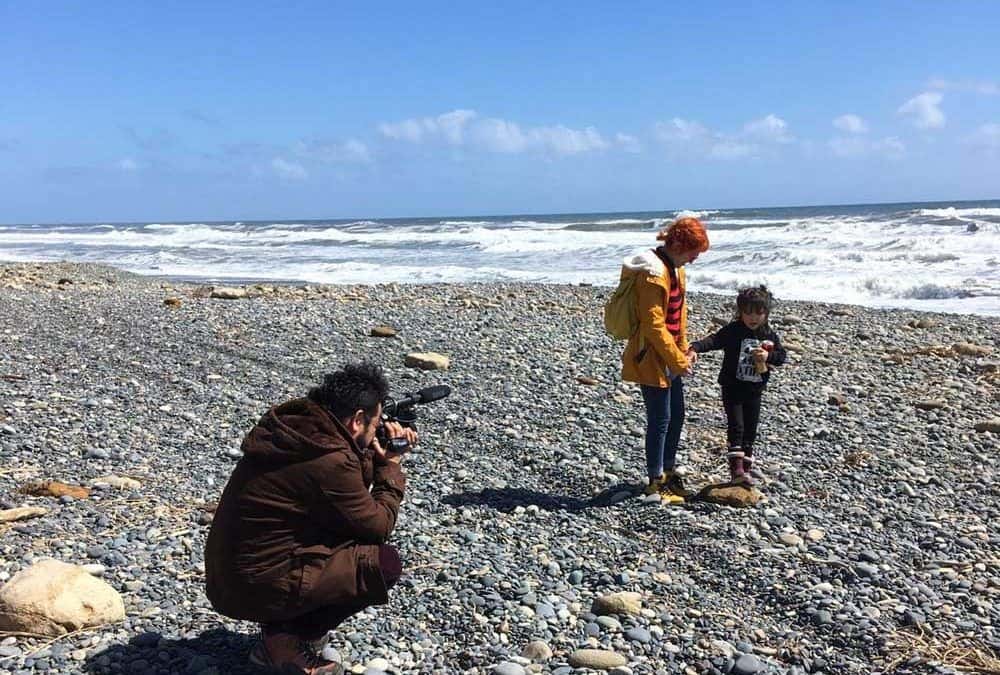 Foto en una playa grande y vacía en un día soleado en el sur de Chile, donde aparece una mujer de su mano con su hija caminando por la orilla de la playa, y al lado una persona que va grabando todo.
