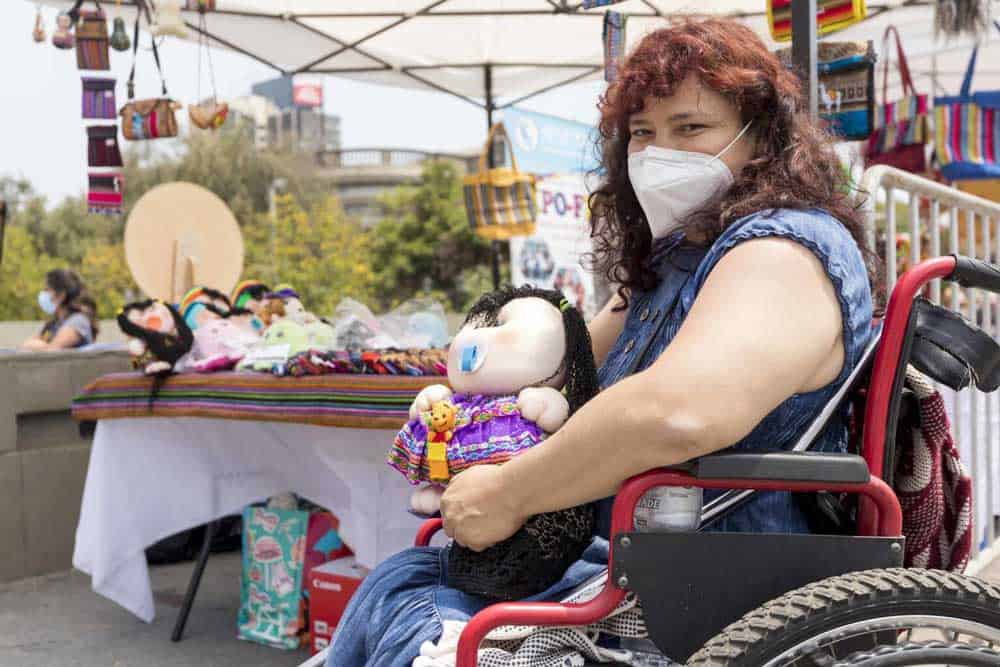 Foto en una feria artesanal donde aparece una mujer en silla de ruedas con una muñeca en la mano y atrás una mesa con más muñecas hechas por ella.