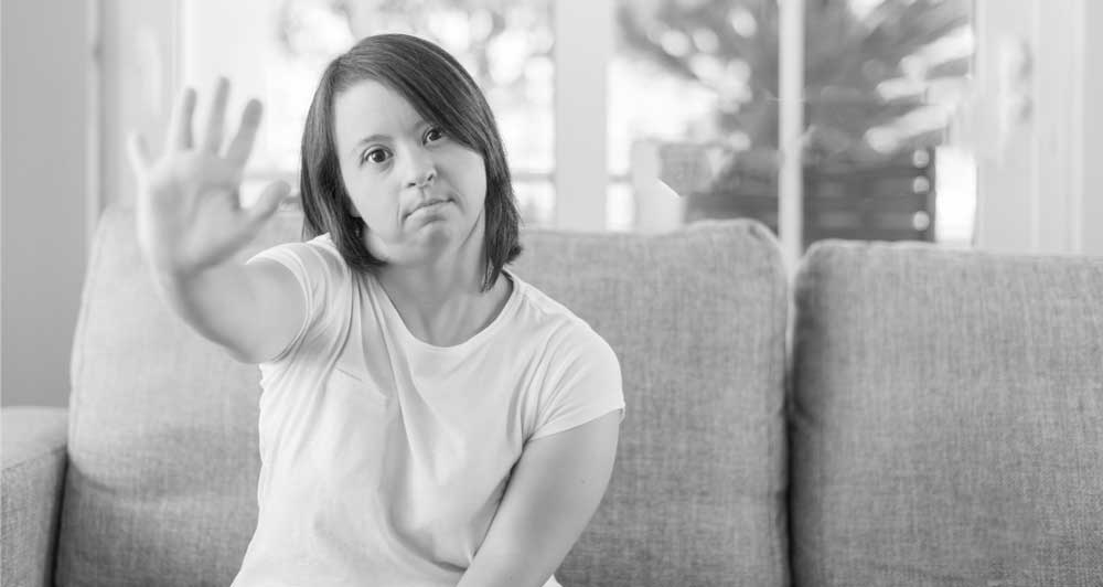 Foto de una mujer con síndrome de down sentada en el sillón de una casa y pone su mano derecha abierta hacia adelante en señal de detener algo.
