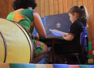 Foto en una sala de clases donde aparece una profesora junto a una alumna pequeña sentadas frente a un computador.